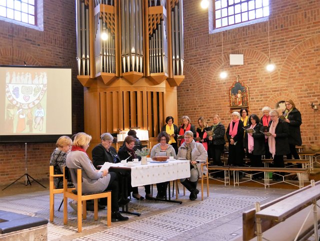 Einige der Frauen des ökumenischen Vorbereitungsteams sitzen am gedeckten Kaffeetisch, dahinter der Chor Intermezzo unter Leitung von Ursula Papon, der mit slowenischen Liedern den Gottesdienst musikalisch begleitete, und links das Titelbild des Weltgebetstag 2019 zu Slowenien, per Beamer auf die Leinwand projeziert.